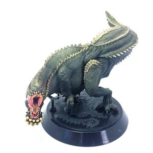 May include: A green and gold dinosaur figurine with a large open mouth and sharp teeth. The dinosaur is standing on a black base with a detailed design.
