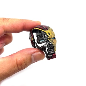 May include: A miniature Iron Man helmet, held between fingers, showcasing a detailed design. The helmet features a metallic gold and red color scheme with intricate mechanical details, including blue eye lenses. The overall aesthetic suggests a collectible or display item.