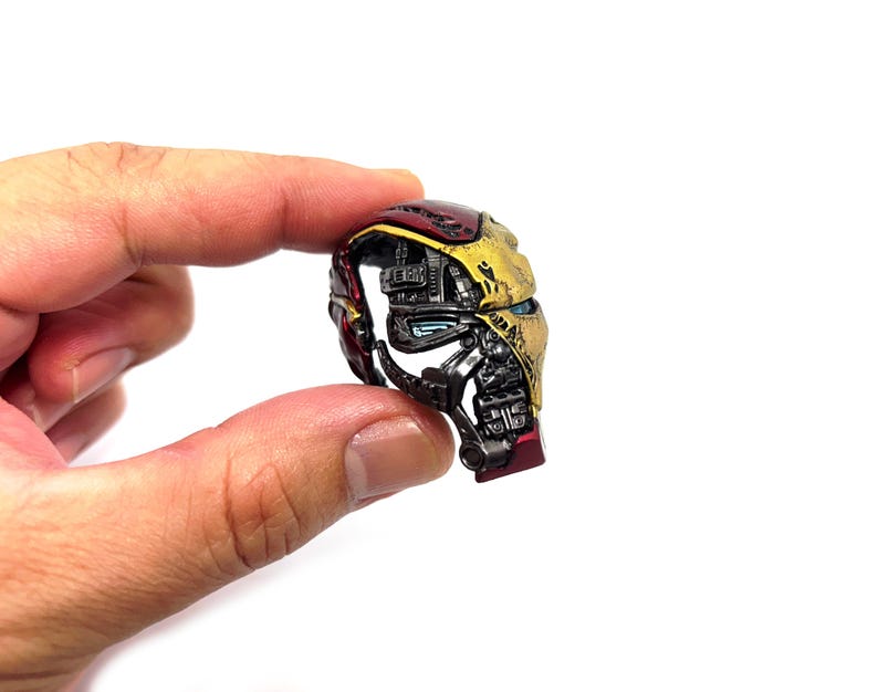 May include: A miniature Iron Man helmet, held between fingers, showcasing a detailed design. The helmet features a metallic gold and red color scheme with intricate mechanical details, including blue eye lenses. The overall aesthetic suggests a collectible or display item.