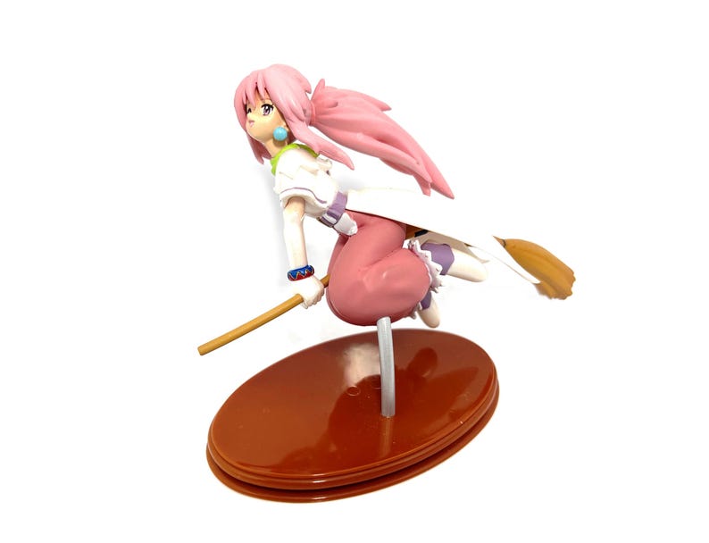 Op de afbeelding: Een beeldje van een vrouwelijk anime-personage met roze haar, gekleed in een witte en paarse outfit, rijdend op een bezem. Het beeldje staat op een bruine ovale basis en heeft een gedetailleerd ontwerp. Het personage staat in een dynamische pose.