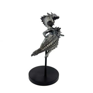 May include: A silver-colored metallic dragon figurine featuring intricate scale textures, a long tail, and a raised head with an open mouth. The creature is mounted on a thin black vertical rod attached to a circular black base for display.
