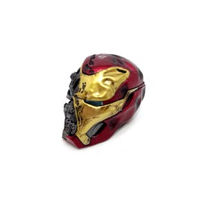 May include: A miniature Iron Man helmet, predominantly red and gold, with battle-damaged detailing. The helmet's design includes a hinged opening, suggesting it may be a container or a collectible item. The color scheme is a mix of metallic red and gold.