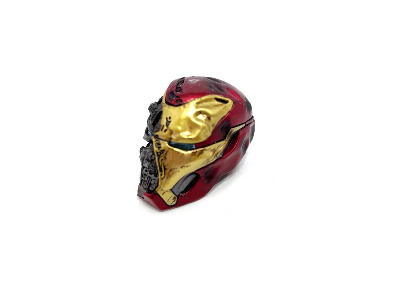 May include: A miniature Iron Man helmet, predominantly red and gold, with battle-damaged detailing. The helmet's design includes a hinged opening, suggesting it may be a container or a collectible item. The color scheme is a mix of metallic red and gold.