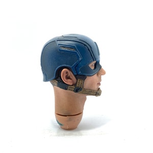 Op de afbeelding: Een blauw en zilver Captain America helm, een verzamelobject. De helm is een gedetailleerde replica, met een textuurontwerp en riemen. Hoofd en nek zijn ook zichtbaar.
