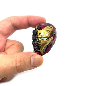 May include: A miniature Iron Man helmet, predominantly red and gold, with intricate detailing. The helmet's design includes visible mechanical components and a partially open faceplate, showcasing the character's iconic look. The helmet is held against a white background.
