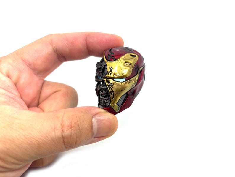 May include: A miniature Iron Man helmet, predominantly red and gold, with intricate detailing. The helmet's design includes visible mechanical components and a partially open faceplate, showcasing the character's iconic look. The helmet is held against a white background.