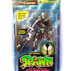 Peut inclure: Une figurine d'action verte et noire de Spawn, un personnage de la série de bandes dessinées du même nom. La figurine tient une tronçonneuse et a un bras cybernétique. La figurine est emballée dans une boîte en plastique avec un fond bleu et blanc. L'emballage présente le logo Spawn et le texte "Deluxe Edition Ultra-Action Figures".