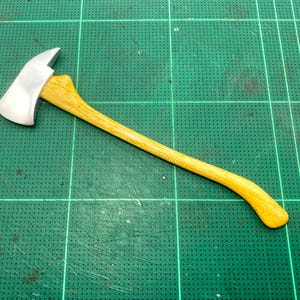 以下が含まれることがあります： 銀色の斧で、木製の柄がついています。柄は薄い茶色で、わずかに曲がっています。斧は緑色のカッティングマットの上に置かれています。