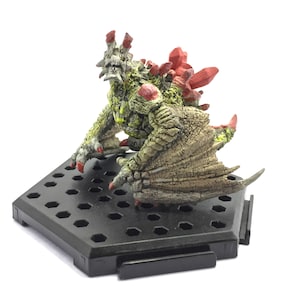 Op de afbeelding: Een groen en rood monsterfiguur met een zwarte zeshoekige basis. Het monster heeft een stekelige rug en vleugels.