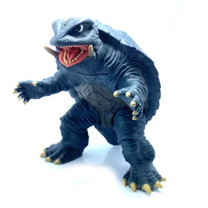 May include: A dark blue, reptilian monster figure with a turtle-like shell, open mouth, and yellow claws. The figure is posed with arms outstretched, showcasing its menacing appearance. The toy is set against a white background.