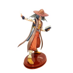 May include: A detailed figurine of a character in a brown hat, orange pants, and a brown and tan vest. The figure is holding a small book and is standing on a red, circular base. The figure has blue hair and is gesturing with one arm.