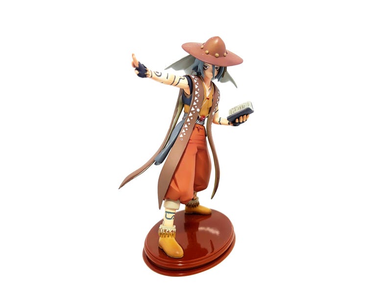 May include: A detailed figurine of a character in a brown hat, orange pants, and a brown and tan vest. The figure is holding a small book and is standing on a red, circular base. The figure has blue hair and is gesturing with one arm.