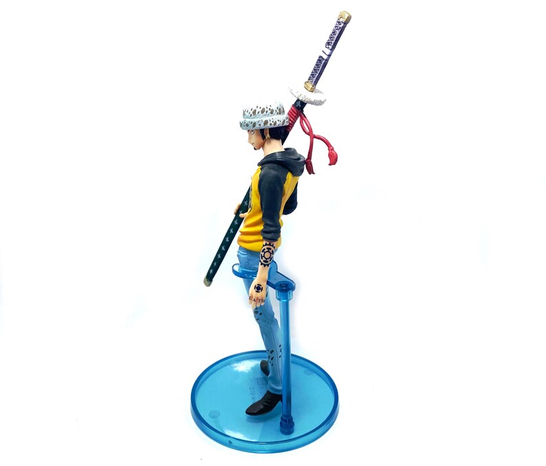 Pu&ograve; includere: Statuetta di un personaggio della serie anime One Piece. La figura indossa una giacca gialla e nera, jeans blu e un cappello bianco con pois neri. Tiene una katana ed &egrave; montata su un supporto blu.