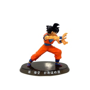 May include: A figurine of a character in a dynamic pose, wearing an orange jumpsuit with blue accents. The figure has black hair and is on a black base with Japanese text. The character is in a fighting stance.