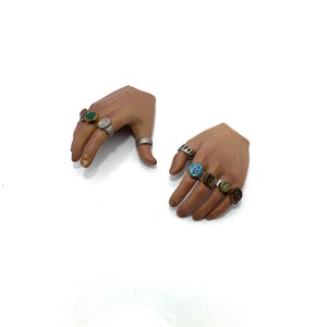 Puede incluir: Dos manos de pl&aacute;stico con anillos en cada dedo. Los anillos son de plata con piedras de diferentes colores, incluyendo verde, azul y marr&oacute;n.