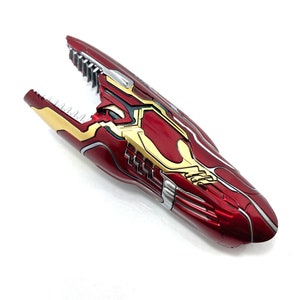 May include: A red and gold gauntlet prop with a metallic finish. The gauntlet features intricate detailing, including silver accents and a claw-like design at the end. The prop is likely inspired by a superhero or science fiction theme.