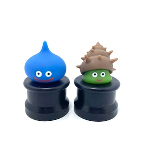 Puede incluir: Dos pequeñas figuritas azules y verdes con caras sonrientes sobre bases negras. La figurita azul tiene forma de lágrima y la figurita verde tiene forma de concha.