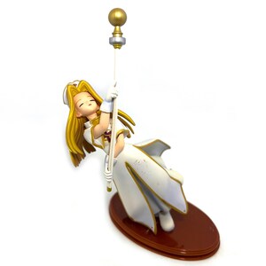 May include: A figurine of a character with long blonde hair, wearing a white and gold outfit, holding onto a staff. The figurine is on a brown oval base. The staff has a gold ball at the top.