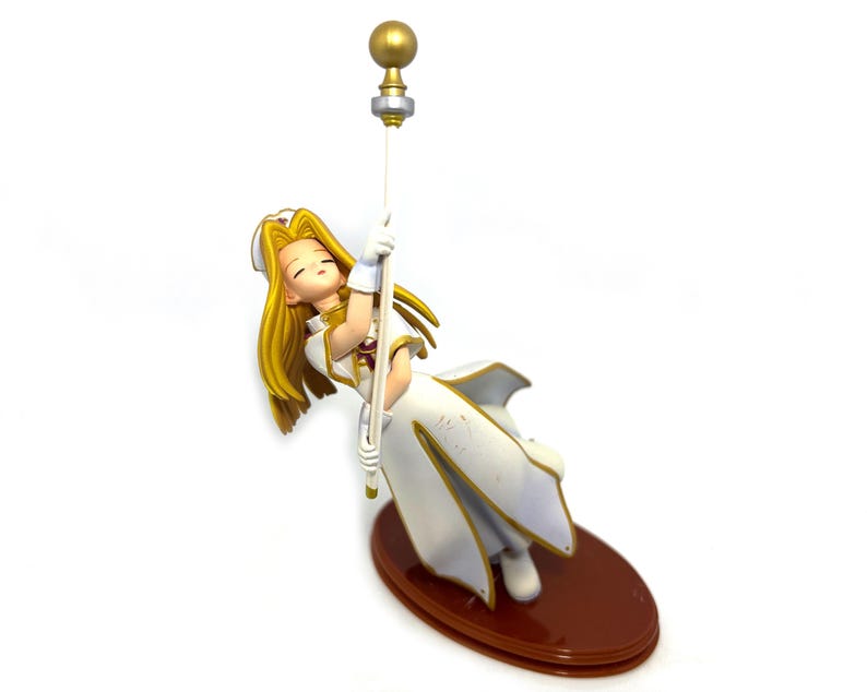 May include: A figurine of a character with long blonde hair, wearing a white and gold outfit, holding onto a staff. The figurine is on a brown oval base. The staff has a gold ball at the top.
