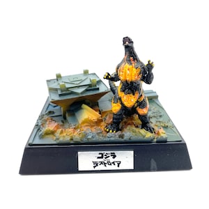 Könnte beinhalten: Eine schwarze und orangefarbene Godzilla-Figur mit Flammen auf dem Körper, die auf einer zerstörten Stadtbasis steht. Die Basis hat eine schwarze Plakette mit japanischem Text, der "Godzilla vs. SpaceGodzilla" lautet.