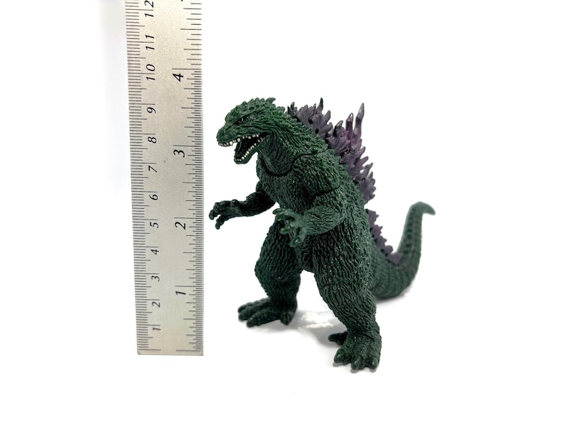 K&ouml;nnte beinhalten: Eine gr&uuml;ne Godzilla-Figur mit lila R&uuml;ckenflossen, etwa 10 cm hoch, steht neben einem Lineal. Das Spielzeug hat einen ge&ouml;ffneten Mund und steht auf einer wei&szlig;en Oberfl&auml;che.