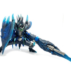 May include: A detailed action figure of a warrior in a crouching pose, wielding a large shield and a lance. The figure is predominantly dark blue and silver, with accents of light blue and green. The shield and lance have intricate, textured designs.