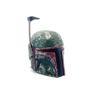 May include: A weathered green and red Boba Fett helmet with a black visor and a silver antenna. The helmet is made of plastic and has a distressed look.