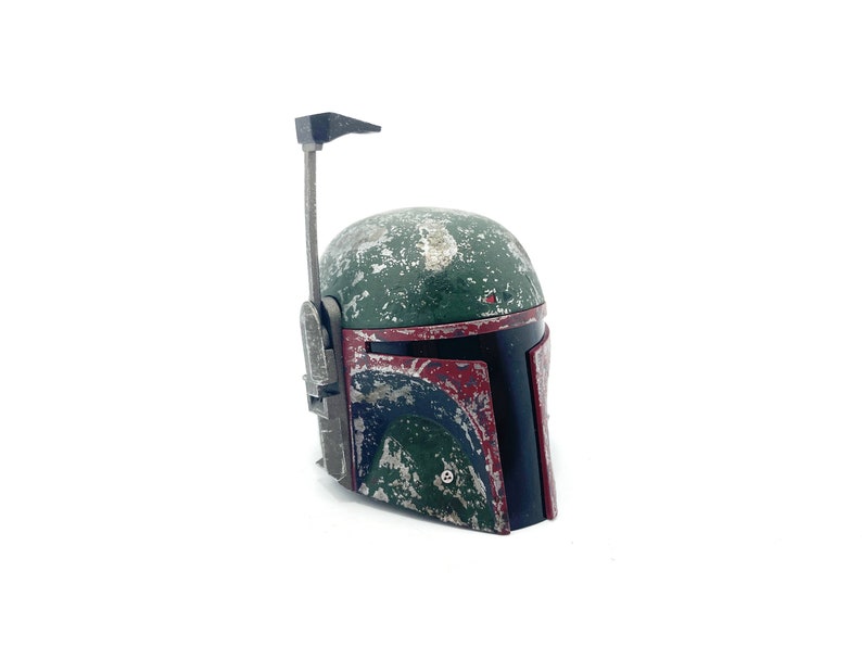 May include: A weathered green and red Boba Fett helmet with a black visor and a silver antenna. The helmet is made of plastic and has a distressed look.