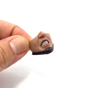 以下が含まれることがあります： ミニチュアアクションフィギュアの頭部の詳細なクローズアップ。歯を食いしばったしかめっ面が特徴です。フィギュアの顔は日焼けしており、口と顎の周りに黒いマスクが付いています。白い背景に、手で頭部が持たれています。