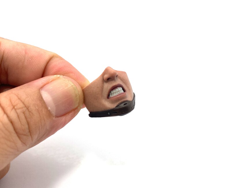 以下が含まれることがあります： ミニチュアアクションフィギュアの頭部の詳細なクローズアップ。歯を食いしばったしかめっ面が特徴です。フィギュアの顔は日焼けしており、口と顎の周りに黒いマスクが付いています。白い背景に、手で頭部が持たれています。