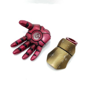 May include: A red and gold plastic Iron Man hand and forearm. The hand is open and the forearm is closed. The hand has a black detail around the fingers and a black circle in the palm. The forearm is gold with a red ring at the top.