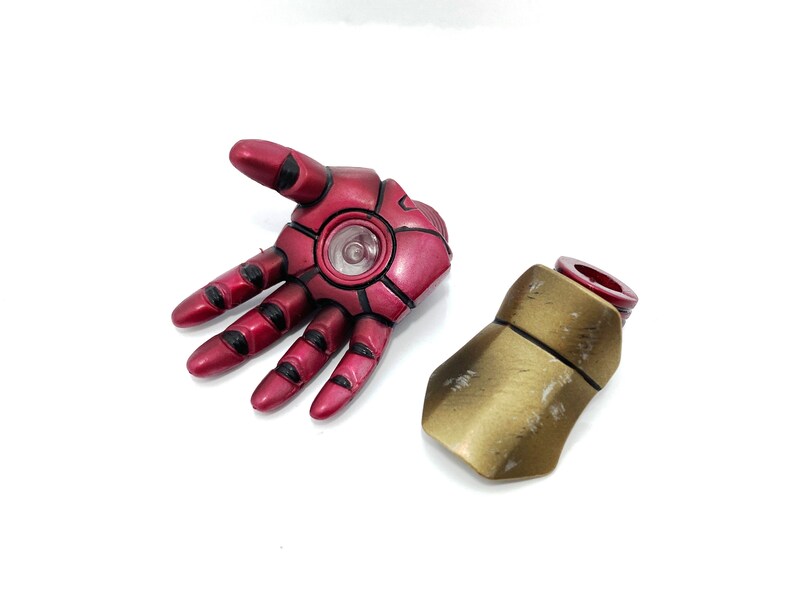 May include: A red and gold plastic Iron Man hand and forearm. The hand is open and the forearm is closed. The hand has a black detail around the fingers and a black circle in the palm. The forearm is gold with a red ring at the top.