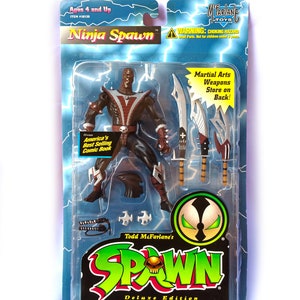 May include: A plastic package containing a Ninja Spawn action figure from Todd McFarlane's Spawn Deluxe Edition Ultra-Action Figures. The figure is black and red with a skull on its chest. The figure is holding a sword and has a variety of weapons in the package. The package is blue and green with the words "Ninja Spawn" and "Spawn" on it.