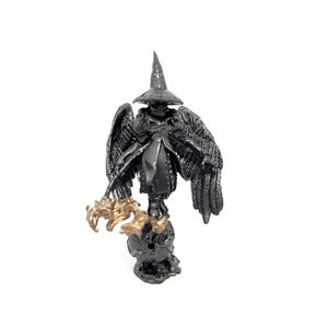 Peut inclure: Figurine sombre et détaillée d'une figure ailée portant un chapeau pointu et une robe. La figurine a des ailes déployées et tient un bâton. La figurine est principalement noire avec des accents dorés et repose sur une base texturée.