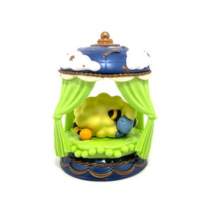 May include: A small figurine featuring a yellow sheep with black and yellow striped legs sleeping on a green circular bed. The bed is enclosed by a blue canopy with white clouds and gold stars, supported by four light green curtains.