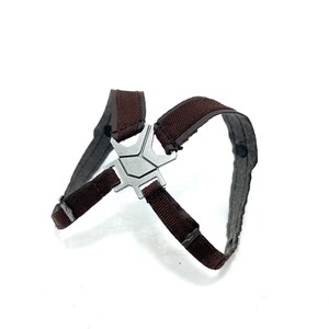 May include: A brown and gray harness with a silver-colored metal clasp. The harness has a cross-like design with adjustable straps. The background is plain white.