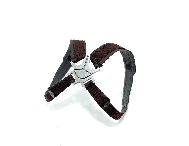 May include: A brown and gray harness with a silver-colored metal clasp. The harness has a cross-like design with adjustable straps. The background is plain white.