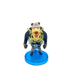 May include: A small plastic figurine of a character from the anime One Piece. The character is wearing a black and white jacket with a yellow symbol on the chest. The figurine is standing on a blue base with the word "Kuma" written on it.