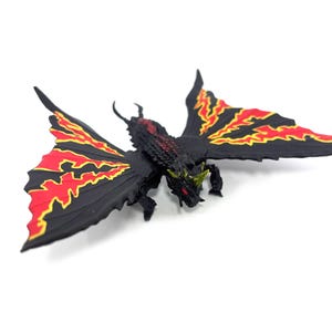 May include: A black creature figurine with large, spread wings featuring jagged red and yellow patterns. The figure has a textured body, a horned head with red eyes, and small clawed limbs. It is a detailed, multi-colored fantasy monster model.