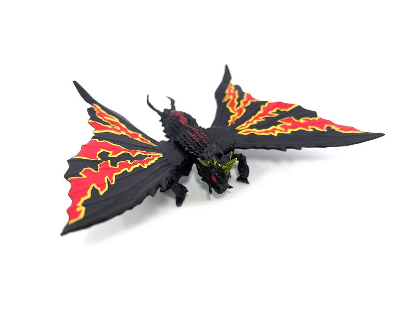 May include: A black creature figurine with large, spread wings featuring jagged red and yellow patterns. The figure has a textured body, a horned head with red eyes, and small clawed limbs. It is a detailed, multi-colored fantasy monster model.