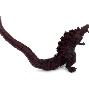 May include: A dark red Godzilla figurine with a long, segmented tail and dorsal fins. The figure is posed with its mouth open, showcasing its teeth. The toy is set against a white background.