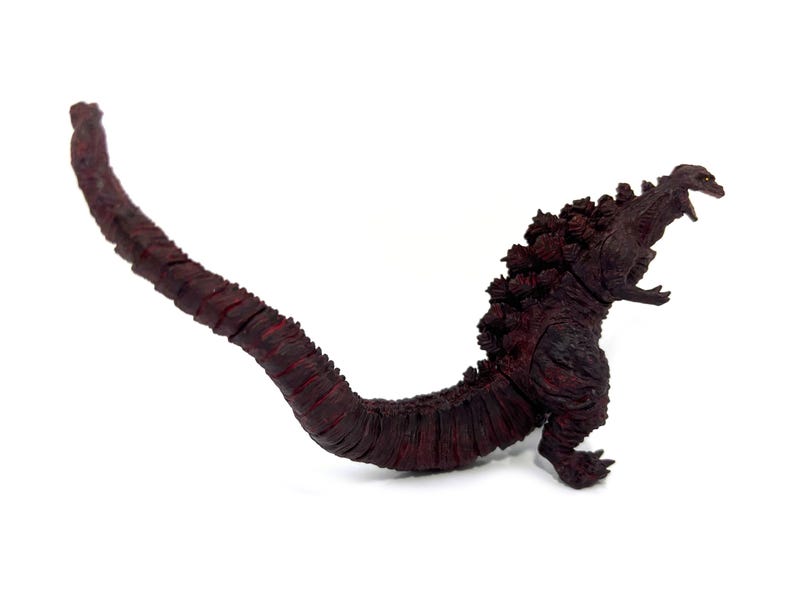May include: A dark red Godzilla figurine with a long, segmented tail and dorsal fins. The figure is posed with its mouth open, showcasing its teeth. The toy is set against a white background.
