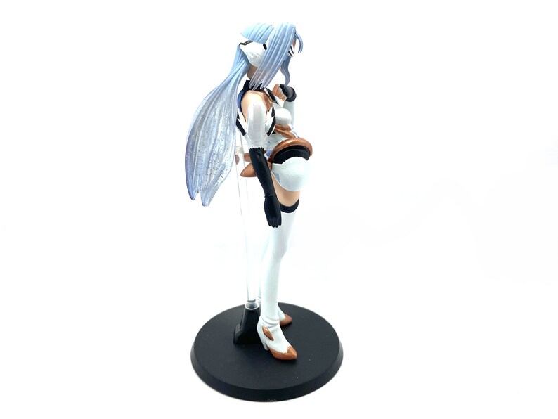 May include: A detailed figurine of a character with long, light blue hair and a white and brown outfit. The figure stands on a black circular base with a clear support rod. The figure has a dynamic pose and intricate design.