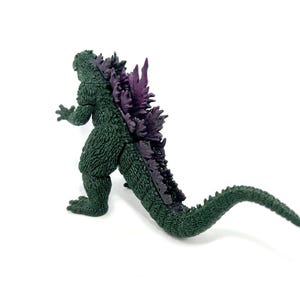 K&ouml;nnte beinhalten: Eine gr&uuml;ne Godzilla-Figur mit lila R&uuml;ckenflossen und einem langen Schwanz. Die Figur ist auf wei&szlig;em Hintergrund platziert und zeigt ihre strukturierte Haut und ihr detailliertes Design. Ein Sammlerst&uuml;ck.