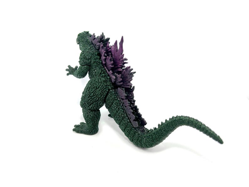 K&ouml;nnte beinhalten: Eine gr&uuml;ne Godzilla-Figur mit lila R&uuml;ckenflossen und einem langen Schwanz. Die Figur ist auf wei&szlig;em Hintergrund platziert und zeigt ihre strukturierte Haut und ihr detailliertes Design. Ein Sammlerst&uuml;ck.