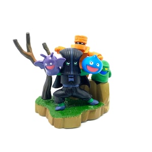 Pode incluir: Um grupo de quatro pequenas figuras de plástico, incluindo uma criatura preta e roxa, uma criatura azul e verde, uma criatura verde e uma criatura laranja, estão em pé sobre uma base verde e marrom. As figuras fazem parte de um conjunto de brinquedos colecionáveis.