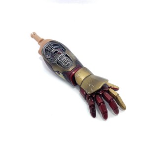 K&ouml;nnte beinhalten: Ein detaillierter Actionfigurenarm mit einem metallischen Gold- und Rot-Design. Der Arm der Figur hat einen realistischen Hautton an der Schulter und ist vor einem wei&szlig;en Hintergrund platziert. Die Hand befindet sich in einer entspannten Position.
