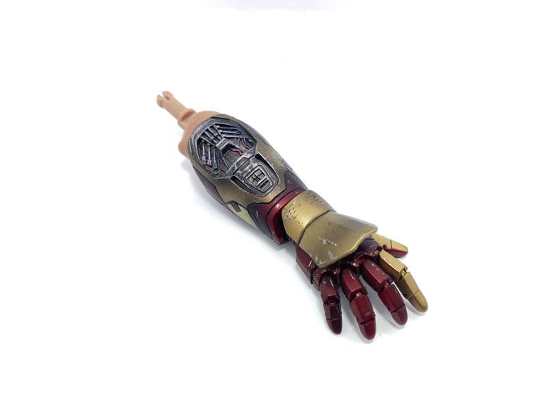 K&ouml;nnte beinhalten: Ein detaillierter Actionfigurenarm mit einem metallischen Gold- und Rot-Design. Der Arm der Figur hat einen realistischen Hautton an der Schulter und ist vor einem wei&szlig;en Hintergrund platziert. Die Hand befindet sich in einer entspannten Position.