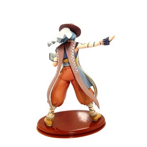 May include: A figurine of a character in a hat and long coat, with orange pants and yellow boots, standing on a red base. The character has blue hair and is holding a small book. The coat is brown with white and blue accents.