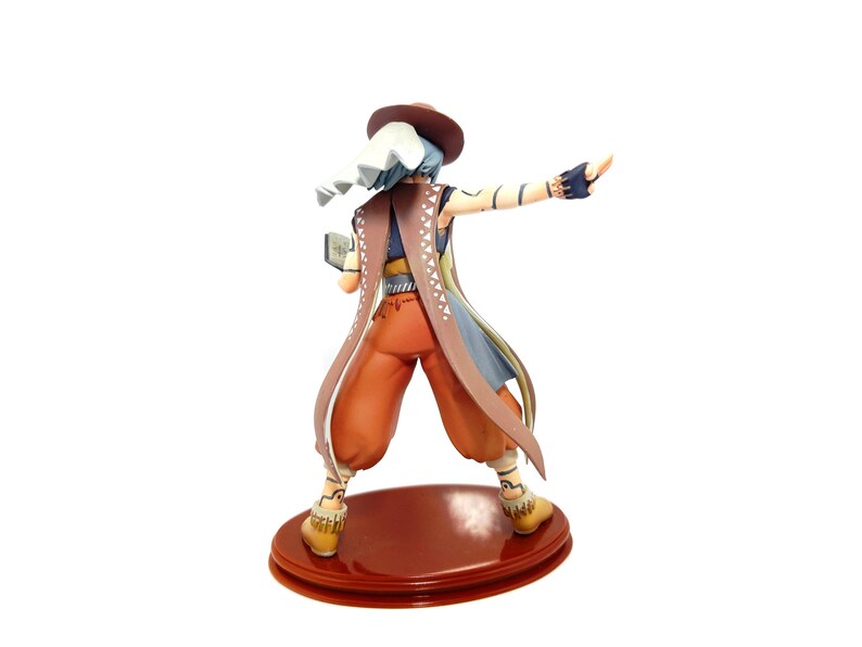May include: A figurine of a character in a hat and long coat, with orange pants and yellow boots, standing on a red base. The character has blue hair and is holding a small book. The coat is brown with white and blue accents.